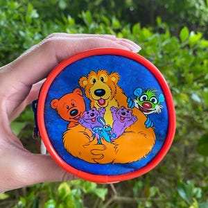 Puede incluir: Una pequeña cartera redonda con borde rojo y fondo azul con una imagen colorida de una familia de osos. La familia de osos es naranja, morada y verde. La familia de osos está rodeada de un fondo azul con un patrón de remolino blanco.