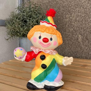 May include: A ceramic clown figurine with a colourful outfit. The clown has orange hair, a red nose, and rosy cheeks. It wears a rainbow-striped outfit with black buttons and a ruffled collar. The clown is holding a star-shaped ball and has a party hat.