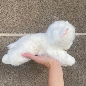 May include: A white plush toy cat with pink ears. The cat is lying on its side with its head tilted up.
