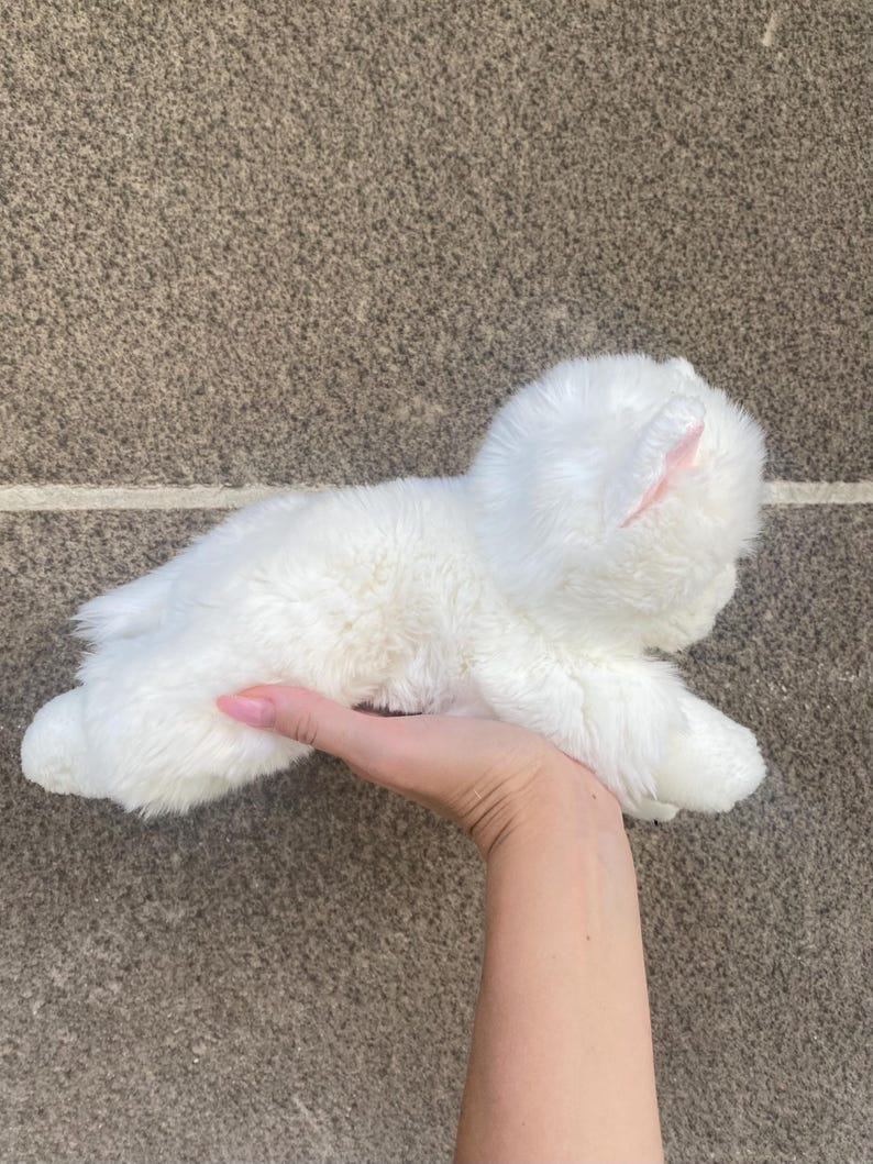 May include: A white plush toy cat with pink ears. The cat is lying on its side with its head tilted up.