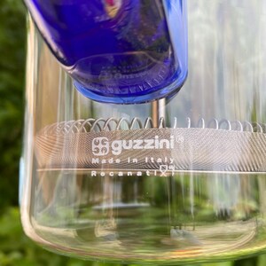 Puede incluir: Una jarra de vidrio transparente con una parte superior azul y un filtro de malla de acero inoxidable. La jarra tiene la marca "guzzini" y "Made in Italy" impresas. Tambi&eacute;n es visible el texto "Recanati".