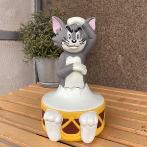 May include: A grey Tom and Jerry cartoon character figurine of Tom the cat playing the drums. Tom is wearing a sailor hat and has a grumpy expression on his face. The drum is yellow with a brown rim and a white drumhead.