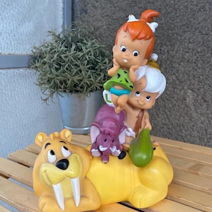 Puede incluir: Una figura amarilla de estilo caricatura con personajes de Los Picapiedra. La figura incluye a Fred y Pebbles, un elefante morado y un tigre dientes de sable amarillo. Los personajes están apilados uno encima del otro. La figura está sobre una superficie de madera.