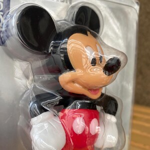 May include: A plastic Mickey Mouse figurine with black ears, a red shirt with a white circle, and a yellow base. The figurine is packaged in a clear plastic bag.