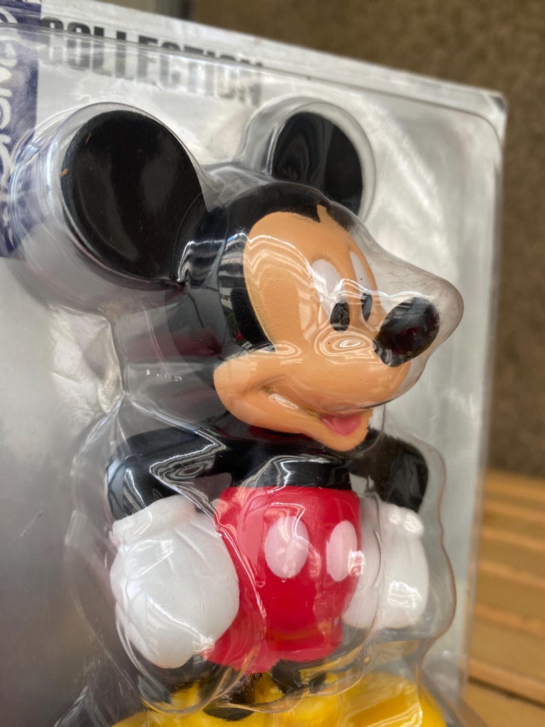 May include: A plastic Mickey Mouse figurine with black ears, a red shirt with a white circle, and a yellow base. The figurine is packaged in a clear plastic bag.