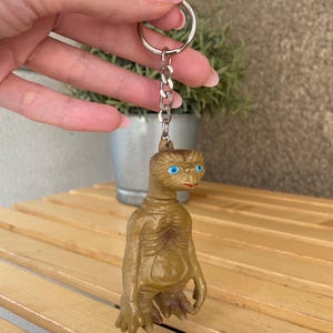 Könnte beinhalten: Ein brauner Plastik-Schlüsselanhänger mit einer kleinen Figur des Außerirdischen E.T. mit großen blauen Augen. Der Schlüsselanhänger hat eine silberne Kette und einen silbernen Ring.