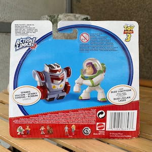 May include: Toy Story 3 Action Links figure set featuring Buzz Lightyear and Sparks Vulcan. Buzz Lightyear is wearing his classic space ranger suit and holding his laser. Sparks Vulcan is a red, white, and blue robot with a large head and a small body. The packaging features the Toy Story 3 logo and the Action Links logo.