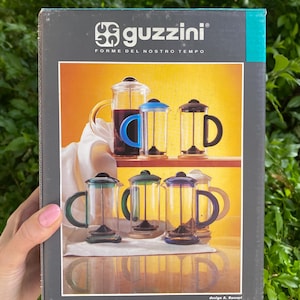 Puede incluir: Una caja de cafetera de prensa francesa Guzzini Orient Express. La caja presenta im&aacute;genes de cafeteras de vidrio con asas y tapas de colores. El texto "guzzini FORME DEL NOSTRO TEMPO" est&aacute; en la parte superior. El texto "Orient Express" est&aacute; en la parte inferior.