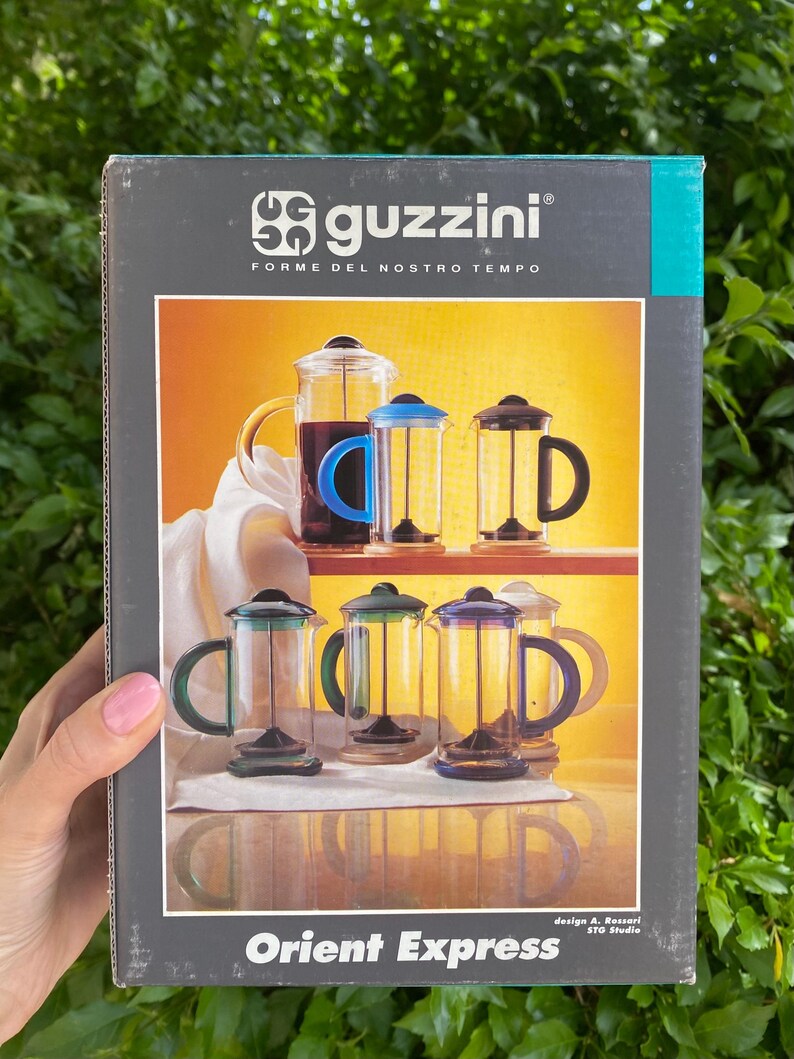Puede incluir: Una caja de cafetera de prensa francesa Guzzini Orient Express. La caja presenta im&aacute;genes de cafeteras de vidrio con asas y tapas de colores. El texto "guzzini FORME DEL NOSTRO TEMPO" est&aacute; en la parte superior. El texto "Orient Express" est&aacute; en la parte inferior.