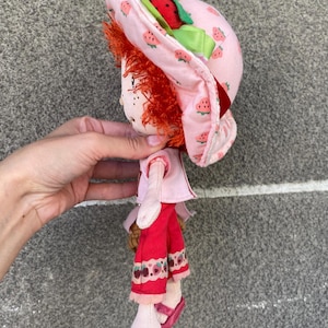 May include: A plush doll with bright red yarn hair wearing a pink sun hat patterned with small strawberries and a green ribbon. The doll features a light pink short-sleeved top, red trousers with a strawberry print hem, and pink slip-on shoes.