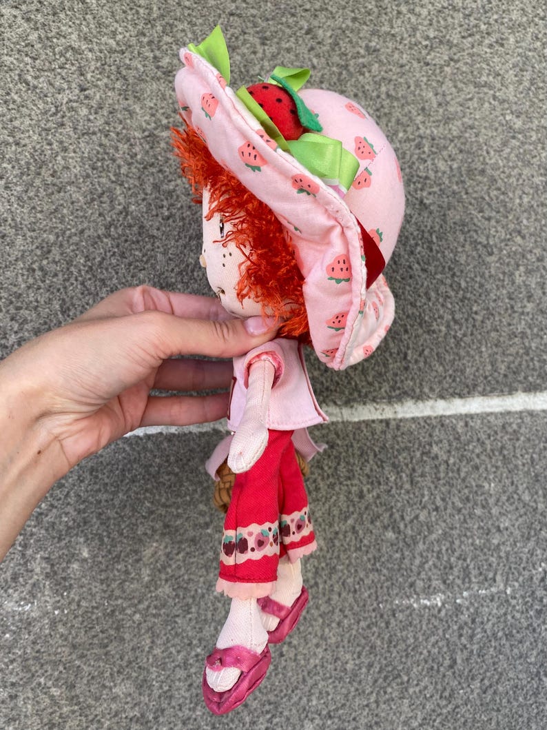 May include: A plush doll with bright red yarn hair wearing a pink sun hat patterned with small strawberries and a green ribbon. The doll features a light pink short-sleeved top, red trousers with a strawberry print hem, and pink slip-on shoes.