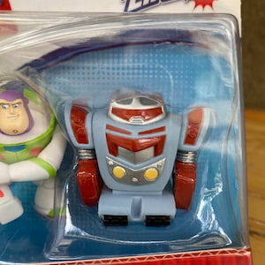May include: A plastic package with a red, white, and blue Action Links Buddy Pack featuring a red, silver, and blue robot toy and a white, blue, and green Buzz Lightyear toy. The package says "Coffret Mini Figurines" and "Buddy Pack".