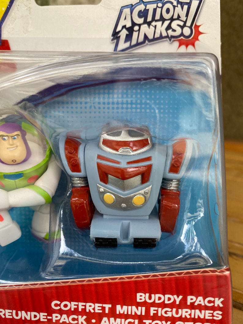 May include: A plastic package with a red, white, and blue Action Links Buddy Pack featuring a red, silver, and blue robot toy and a white, blue, and green Buzz Lightyear toy. The package says "Coffret Mini Figurines" and "Buddy Pack".
