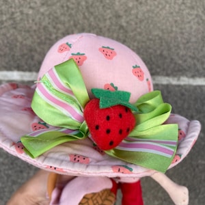 May include: A plush doll wearing a pink sun hat with a strawberry print, accented by a green and white striped ribbon and a three-dimensional red strawberry embellishment. The doll features red trousers and carries a small brown woven basket accessory.