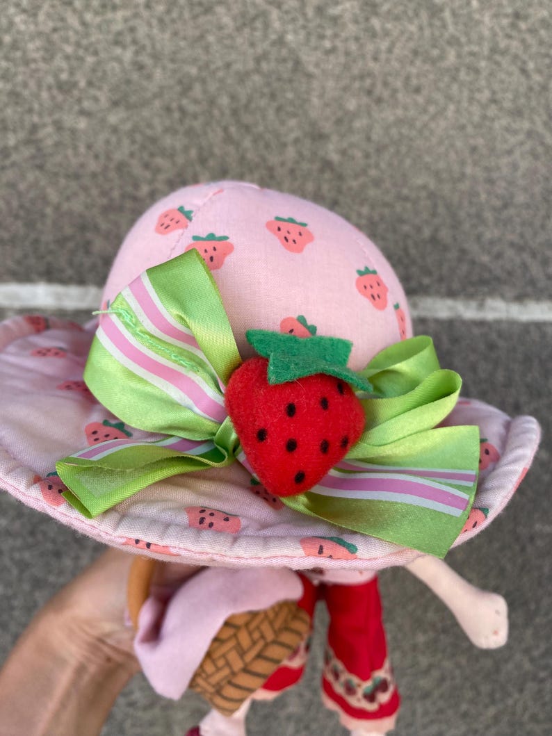 May include: A plush doll wearing a pink sun hat with a strawberry print, accented by a green and white striped ribbon and a three-dimensional red strawberry embellishment. The doll features red trousers and carries a small brown woven basket accessory.
