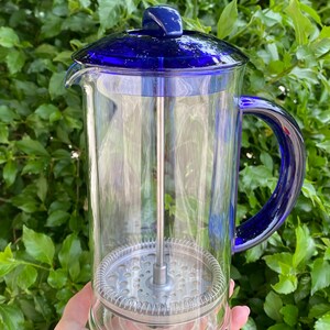 Puede incluir: Una cafetera de prensa francesa de vidrio transparente con una tapa y un asa de color azul vibrante. El interior presenta un filtro y un &eacute;mbolo de acero inoxidable. La cafetera se sostiene contra un fondo de follaje verde.