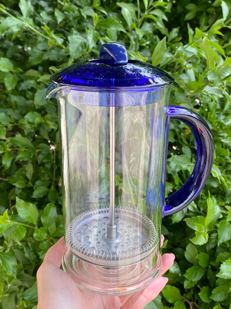 Puede incluir: Una cafetera de prensa francesa de vidrio transparente con una tapa y un asa de color azul vibrante. El interior presenta un filtro y un &eacute;mbolo de acero inoxidable. La cafetera se sostiene contra un fondo de follaje verde.