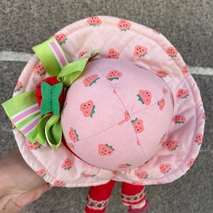 May include: A plush doll wearing a wide-brimmed pink fabric hat with a repeating strawberry pattern. The hat is decorated with a large green and pink striped ribbon bow and a small red felt strawberry accent. The doll has red legs and pink shoes.
