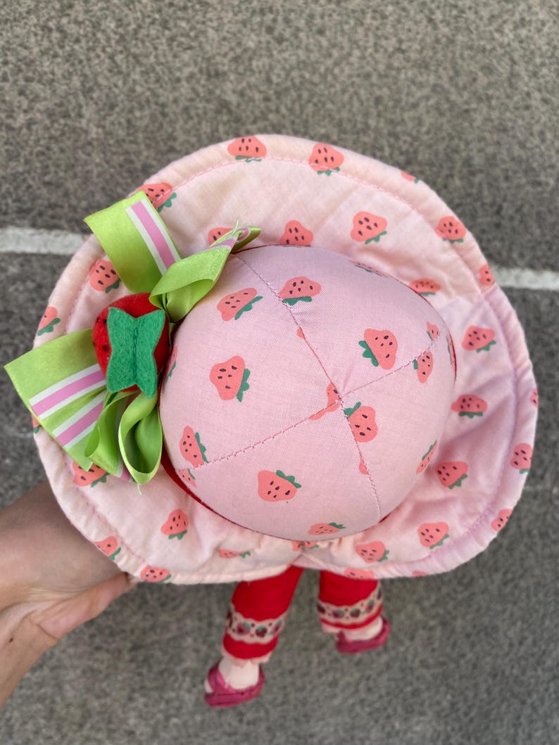 May include: A plush doll wearing a wide-brimmed pink fabric hat with a repeating strawberry pattern. The hat is decorated with a large green and pink striped ribbon bow and a small red felt strawberry accent. The doll has red legs and pink shoes.