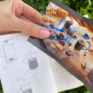Puede incluir: Una persona sostiene un manual de instrucciones y una imagen del producto. El manual muestra diagramas de una cafetera de &eacute;mbolo. La imagen del producto muestra una cafetera de &eacute;mbolo, tazas de caf&eacute; y pasteles. El fondo es verde.