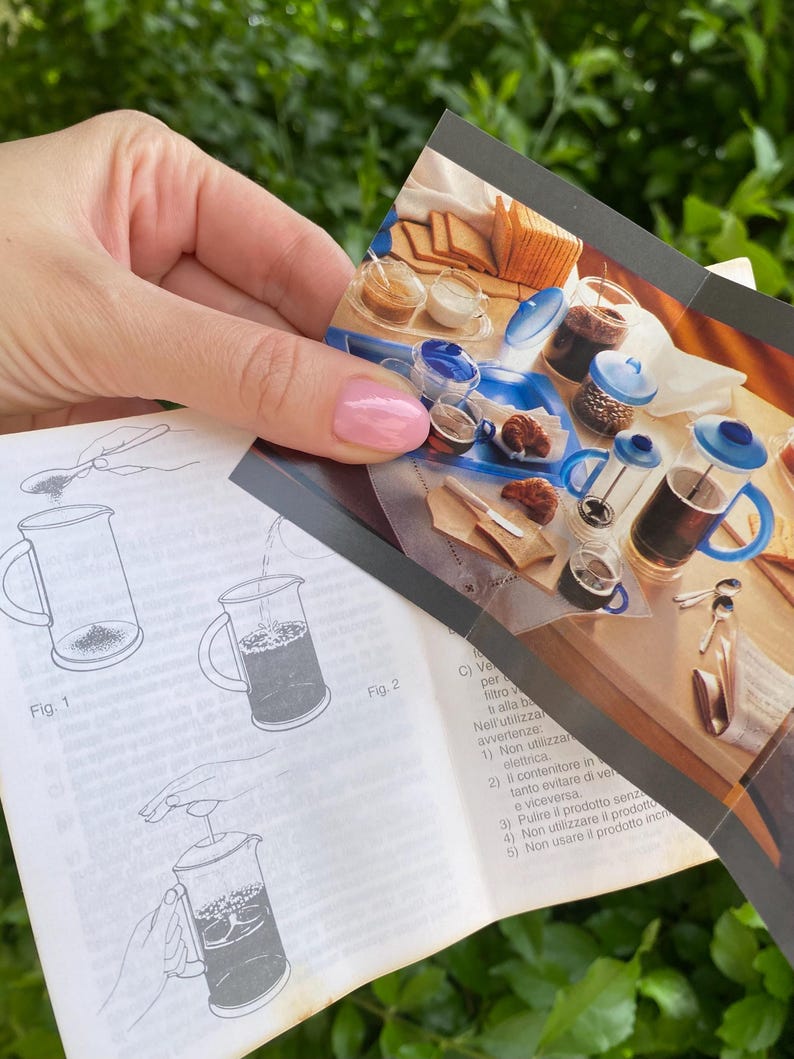 Puede incluir: Una persona sostiene un manual de instrucciones y una imagen del producto. El manual muestra diagramas de una cafetera de &eacute;mbolo. La imagen del producto muestra una cafetera de &eacute;mbolo, tazas de caf&eacute; y pasteles. El fondo es verde.