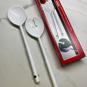 Peut inclure: Un ensemble de deux serveurs à salade en plastique blanc avec des poignées noires. Les serveurs sont conçus avec une cuillère à l'extrémité et une fourchette à l'autre. Les serveurs sont emballés dans une boîte rouge avec le nom de la marque "guzzini" et le nom du produit "CHIMERA" imprimé dessus.