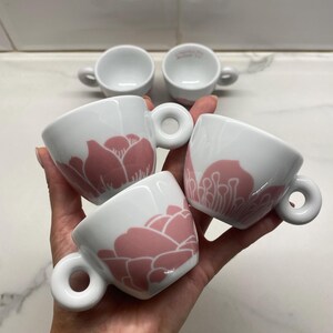 Può includere: Set di quattro tazzine da caffè espresso in ceramica bianca con disegni floreali rosa. Ogni tazza ha un manico arrotondato e un motivo floreale rosa all'esterno. Due tazze sono sullo sfondo, due sono tenute in mano.