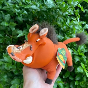 Könnte beinhalten: Ein Plüschtier der Figur Pumbaa aus Der König der Löwen. Das Spielzeug ist orange, braun und hellbraun, mit einem Büschel dunkelbraunen Fells und einem kleinen Schwanz. Es hat ein Etikett mit der Aufschrift "Der König der Löwen".