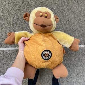 Könnte beinhalten: Ein affenförmiger Rucksack aus Plüsch in Beige- und Brauntönen. Der Affe hat ein lächelndes Gesicht, Schlappohren und einen runden Körper mit einem Reißverschlussfach. Er hat schwarze, verstellbare Träger und ein rundes Logo.