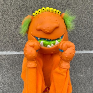 May include: Orange monster hand puppet with green hair, yellow eyes, and a wide grin. The puppet has textured orange hands and a soft orange body. The mouth is green and white. A fun toy for kids.