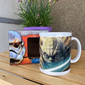 May include: A white ceramic mug featuring a colourful image of Yoda from Star Wars. The mug is in a box with a picture of a Stormtrooper on it. The box is orange and blue with the word "Disney" on it.