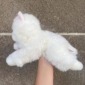 May include: White plush toy shaped like a small llama or alpaca. The toy is being held by a hand.