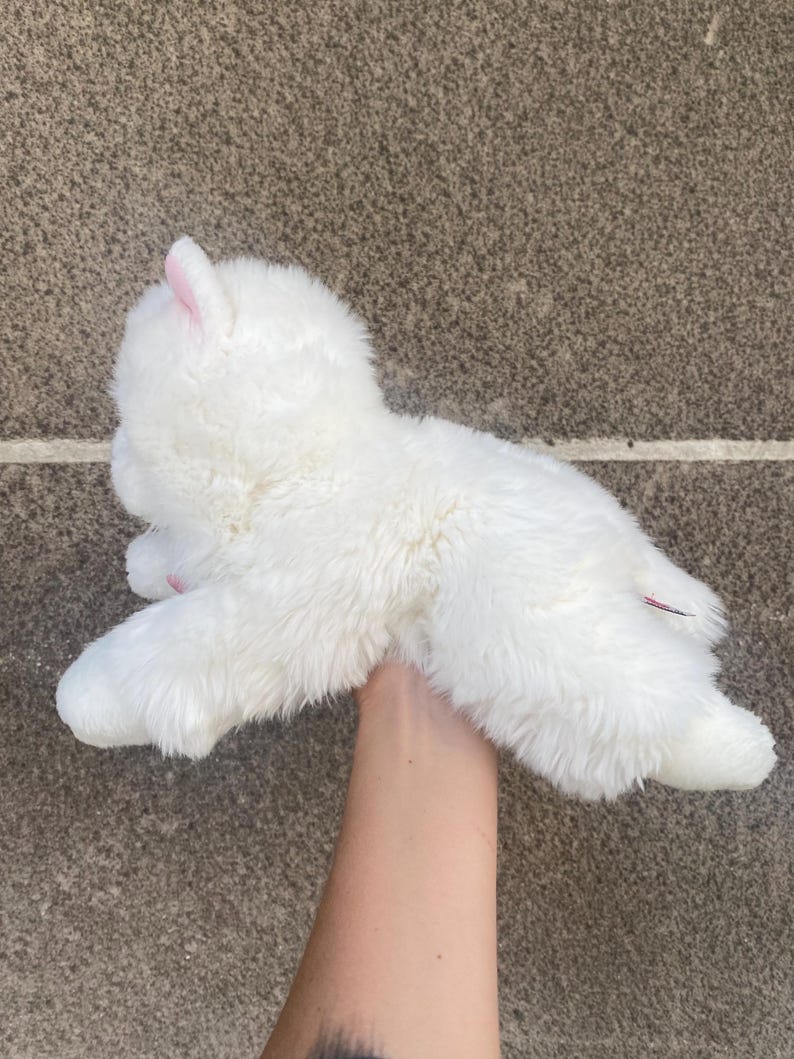May include: White plush toy shaped like a small llama or alpaca. The toy is being held by a hand.