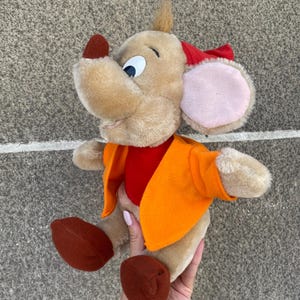 Könnte beinhalten: Ein Plüschmaus-Spielzeug mit beigem Körper, großen Ohren mit rosa Innenseiten und einer roten Schleife. Es trägt eine orangefarbene Jacke über einem roten Hemd und hat braune Füße. Das Spielzeug ist als Zeichentrickfigur gestaltet.