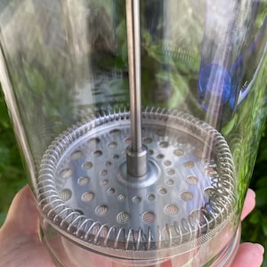 Puede incluir: Una cafetera de prensa francesa de vidrio transparente con un filtro y un &eacute;mbolo de acero inoxidable. La prensa tiene forma cil&iacute;ndrica y est&aacute; dise&ntilde;ada para preparar caf&eacute;. La imagen est&aacute; tomada al aire libre con follaje verde de fondo.