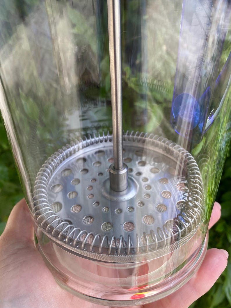 Puede incluir: Una cafetera de prensa francesa de vidrio transparente con un filtro y un &eacute;mbolo de acero inoxidable. La prensa tiene forma cil&iacute;ndrica y est&aacute; dise&ntilde;ada para preparar caf&eacute;. La imagen est&aacute; tomada al aire libre con follaje verde de fondo.