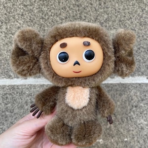 Op de afbeelding: Een bruin pluche speelgoed met grote, pluizige oren en een rond gezicht. Het speelgoed heeft grote zwarte ogen, een kleine rode mond en een lichtbruine vlek op de borst. Het speelgoed wordt in de hand van een persoon gehouden.