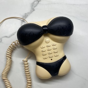 Puede incluir: Teléfono novedoso con forma de torso femenino que lleva un bikini negro. El teléfono tiene un cuerpo beige, sujetador y braguita de bikini negros, y un cable de auricular beige en espiral. Los botones del teclado son visibles.