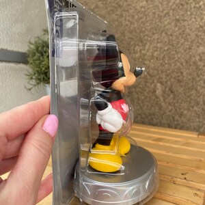 May include: A plastic-wrapped Mickey Mouse figurine with a red shirt and yellow shoes. The figurine is on a silver base with a decorative design. The packaging is labeled "Disney Collectible".