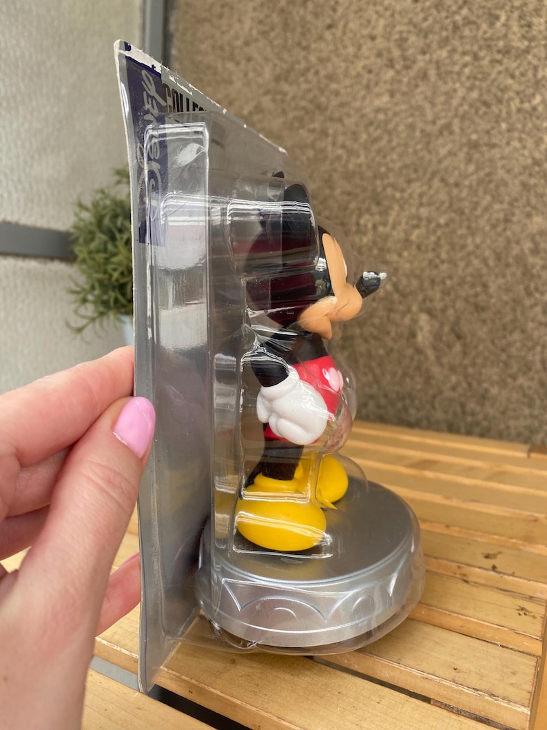 May include: A plastic-wrapped Mickey Mouse figurine with a red shirt and yellow shoes. The figurine is on a silver base with a decorative design. The packaging is labeled "Disney Collectible".