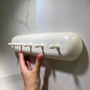 以下が含まれることがあります： 5つの歯ブラシホルダーが付いた白いプラスチック製の歯ブラシホルダー。ホルダーは壁に取り付けられており、歯ブラシを収納するように設計されています。ホルダーは無地の白で、滑らかで丸みを帯びた形状をしています。