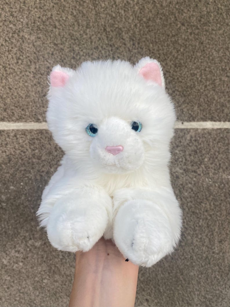 May include: White plush cat toy with pink ears and blue eyes. The cat is sitting up with its paws in front of it.