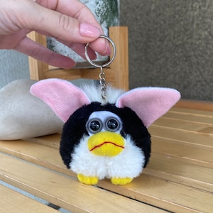 May include: Black and white plush keychain with pink ears and large googly eyes. The keychain is shaped like a cartoon animal with a yellow beak and a small frown.