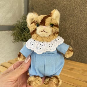 May include: A handmade plush toy cat with brown and tan fur, wearing a light blue outfit with a white lace collar. The cat has green eyes and is approximately 5 inches tall.
