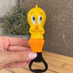 May include: A yellow Tweety Bird bottle opener with blue eyes and an orange beak. The bottle opener has an orange base and a black metal opener. The Tweety Bird figure is a cartoon character.