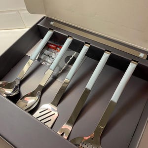 May include: A boxed set of stainless steel kitchen utensils with light blue handles. The set includes a ladle, slotted spoon, spatula, fork, and skimmer. The brand name "guzzini" is visible on the packaging. The utensils are presented in a dark grey box.