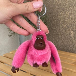 Peut inclure: Un porte-clés orang-outan rose en peluche avec des traits du visage et des pieds marron. Le porte-clés est doté d'un anneau et d'une chaîne en métal argenté. L'orang-outan a une texture duveteuse.