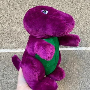 Op de afbeelding: Een pluche paarse dinosaurus speelgoed met een groene buik. Het speelgoed heeft een vriendelijke uitdrukking met witte en blauwe ogen. De dinosaurus is gemaakt van zacht, fuzzy materiaal en heeft een staart en kleine armen.
