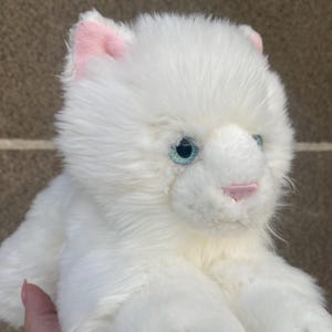 May include: White plush toy cat with blue eyes and pink ears. The cat is lying on its belly with its paws outstretched.