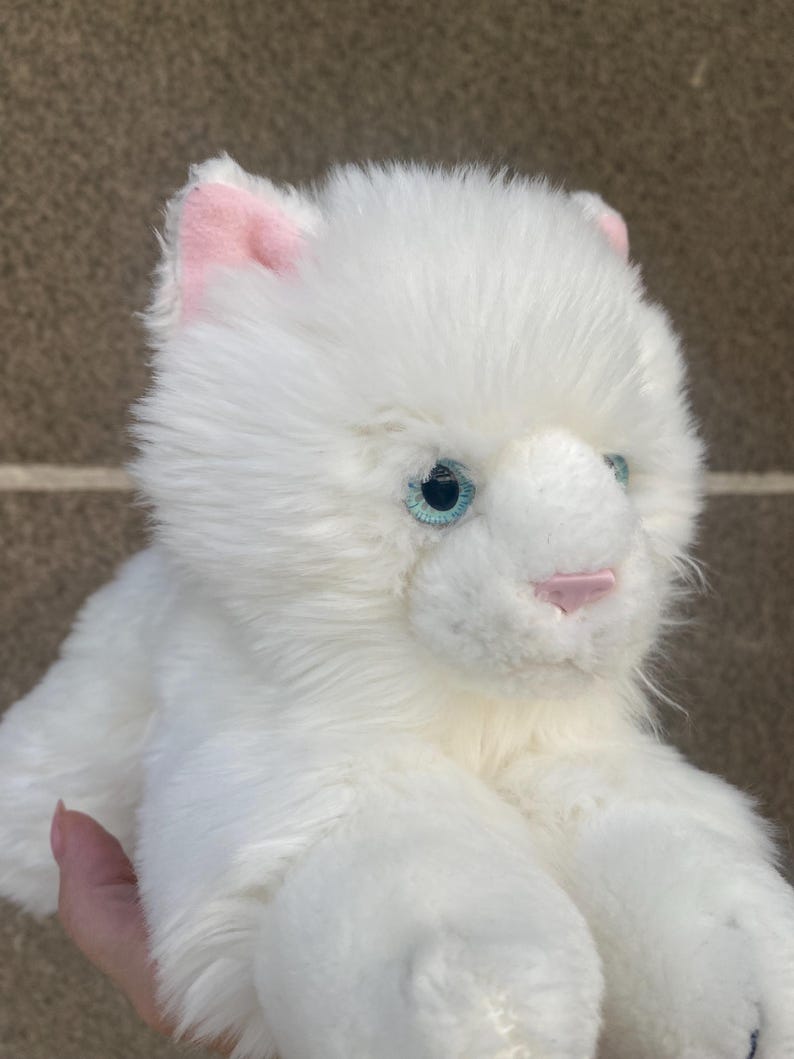 May include: White plush toy cat with blue eyes and pink ears. The cat is lying on its belly with its paws outstretched.